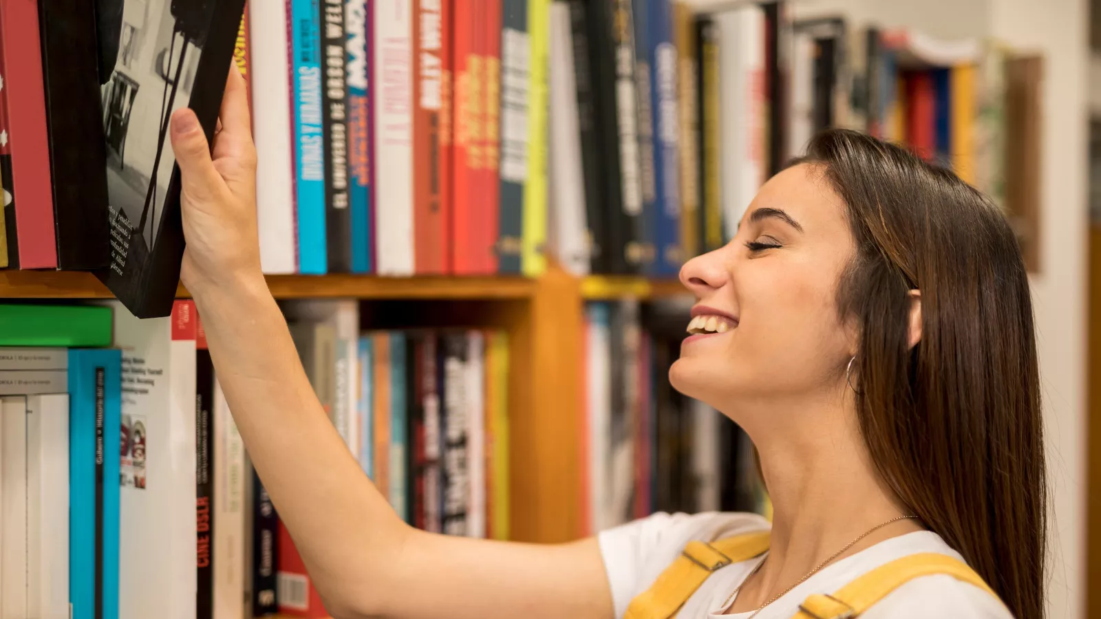 Jovem sorridente de cabelo castanho tira um livro de uma estante cheia de lombadas coloridas em uma livraria, cena ligada ao prêmio para livrarias de rua de São Paulo