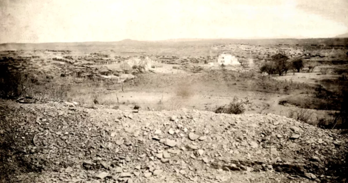 Fotografia histórica em tons de sépia mostra uma paisagem seca e pedregosa, com vegetação esparsa e pequenas construções ao fundo.