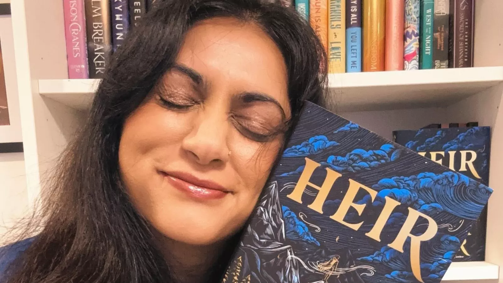 A escritora Sabaa Tahir, de olhos fechados e sorrindo, abraça um exemplar de Heir, primeiro volume de sua duologia, diante de uma estante cheia de livros