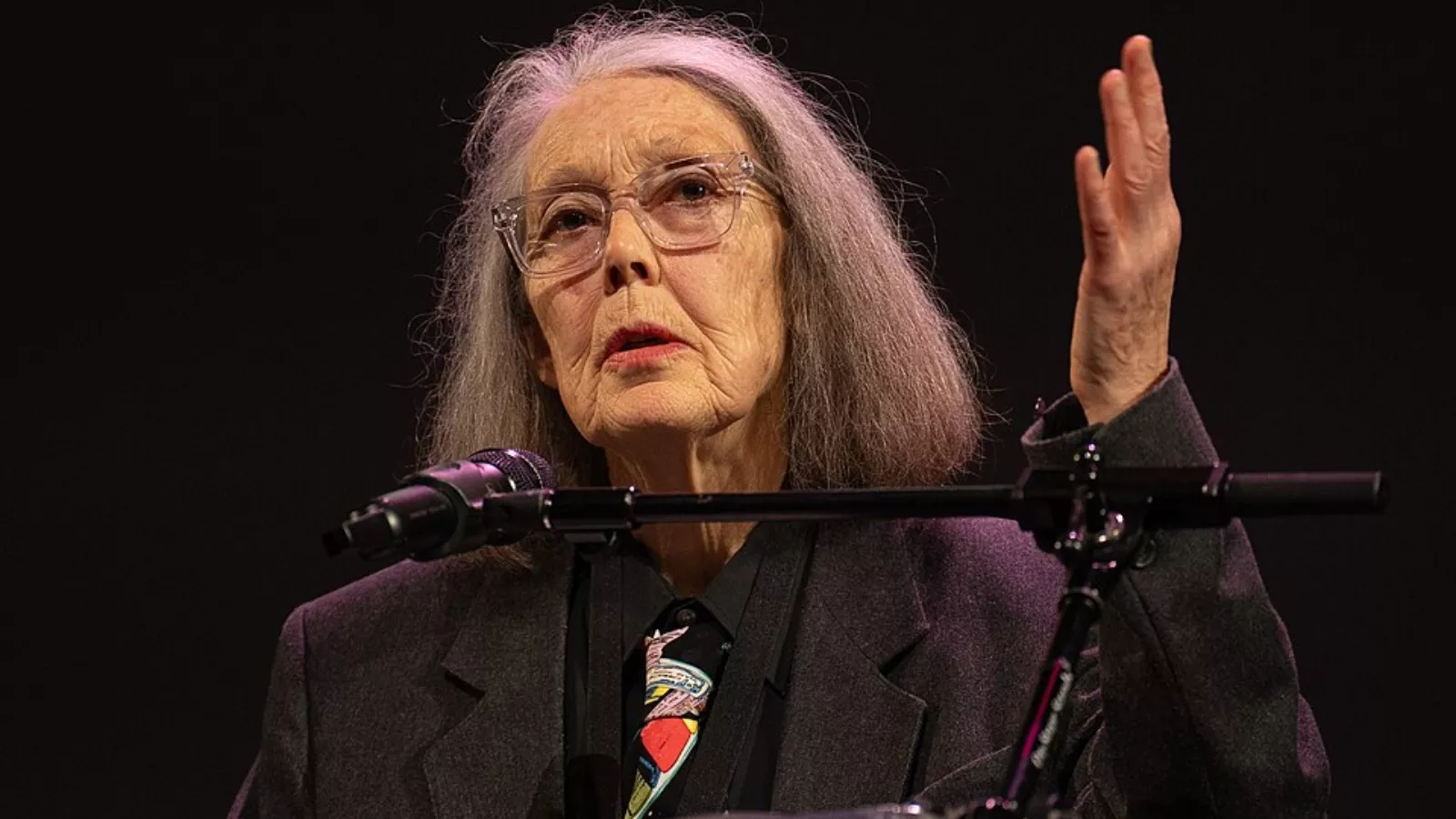 Anne Carson fala ao microfone durante uma apresentação, com uma das mãos levantada.