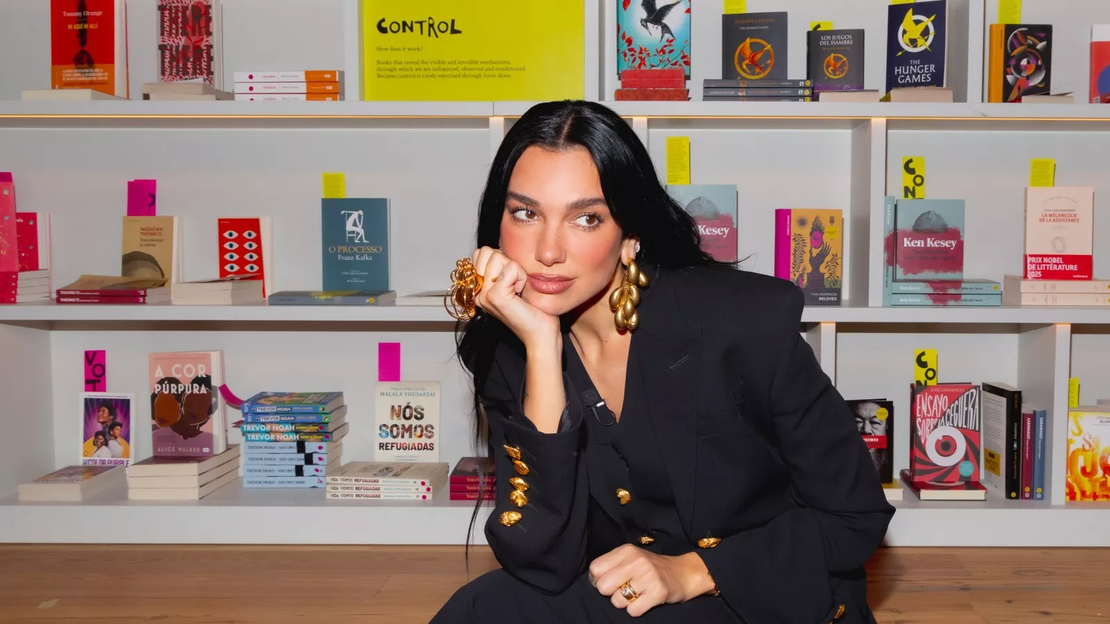 Dua Lipa, de cabelo preto e blazer preto com botões dourados, posa agachada diante das estantes da Manifesto Library, na Livraria Lello, com livros censurados como O Processo
