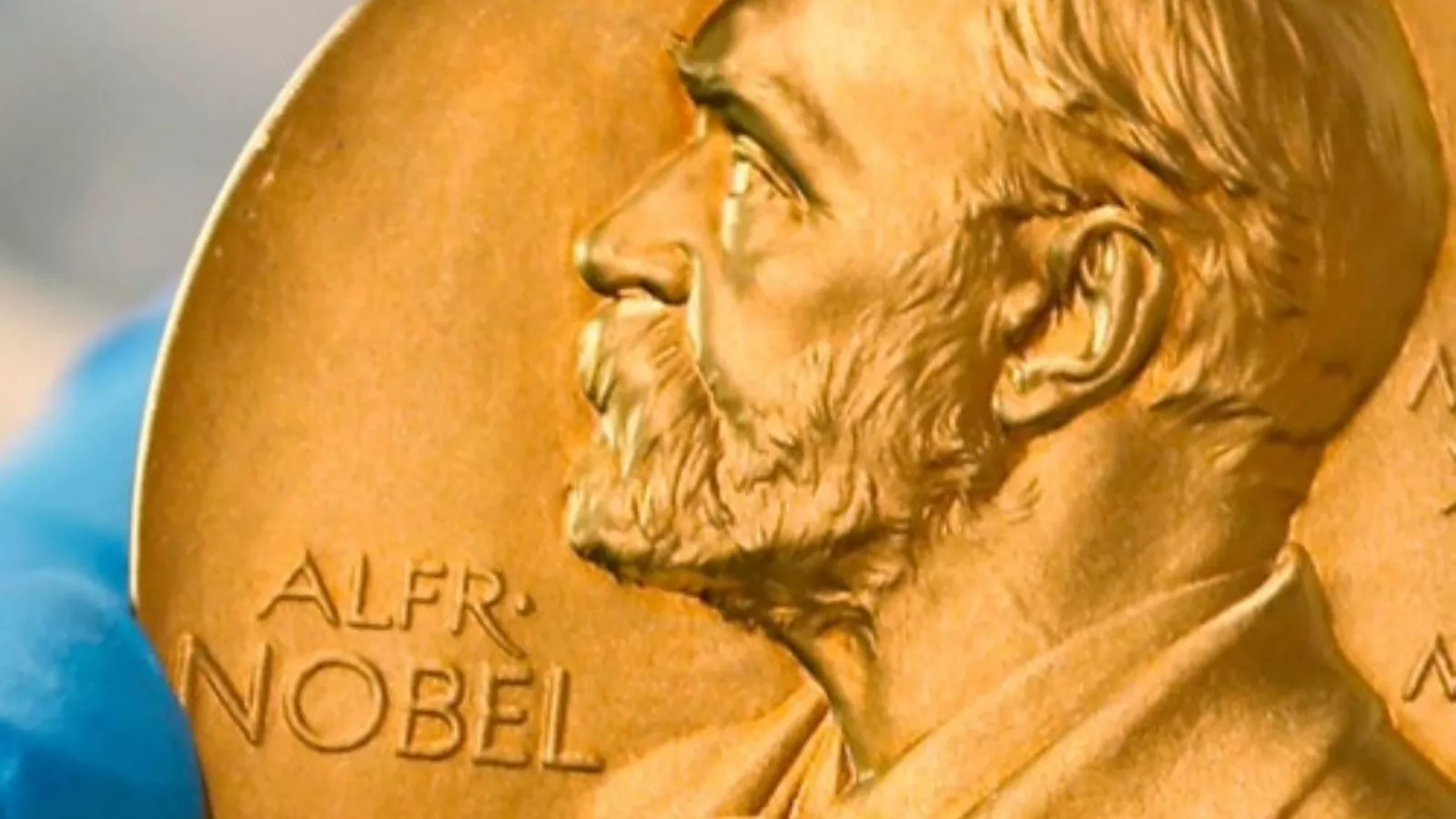 Medalha dourada do Prêmio Nobel em close, com o perfil em relevo de Alfred Nobel e a inscrição Alfr. Nobel, segurada por uma mão de luva azul