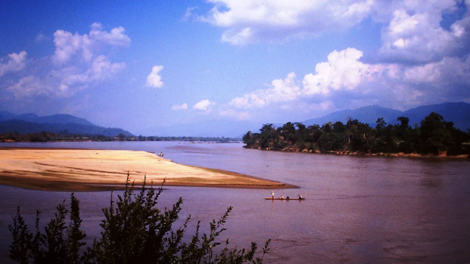 Rio Mekong de águas barrentas, com um barco estreito de pescadores, um banco de areia e margens de mata sob céu azul com nuvens, na região do Triângulo Dourado, em 1985