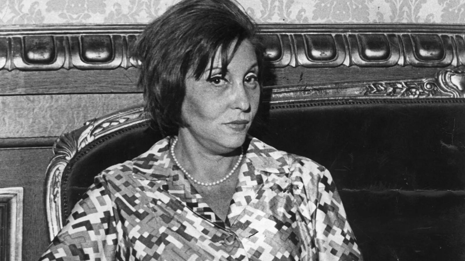 Foto em preto e branco de Clarice Lispector em 1972, sentada num sofá antigo de madeira entalhada. Ela tem cabelo curto com franja, usa colar de pérolas e blusa de estampa geométrica, e olha de lado para a câmera.