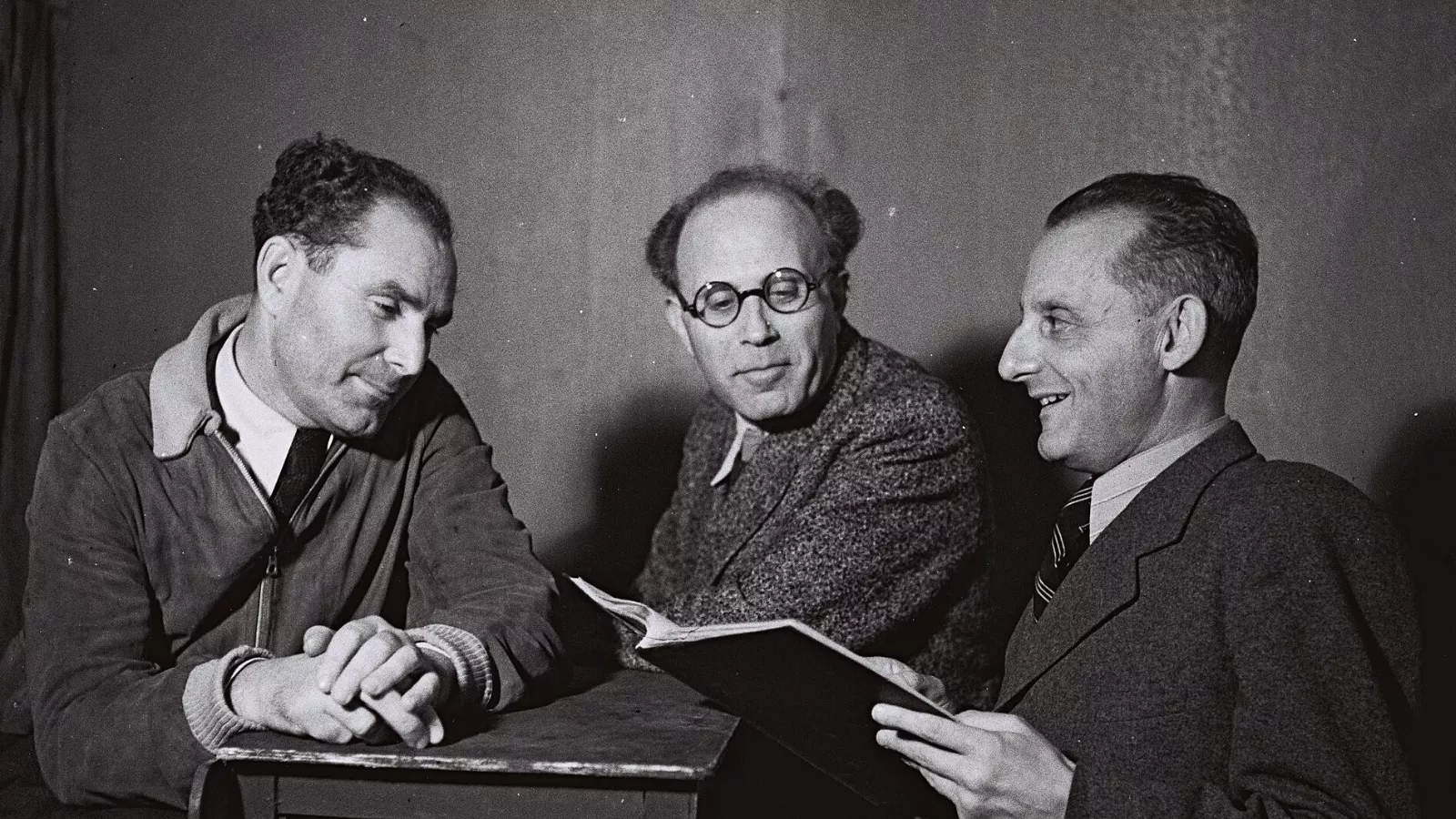 Max Brod, à direita, sorrindo e segurando um livro aberto, ao lado de dois diretores de teatro sentados a uma mesa, em foto em preto e branco de 1942.