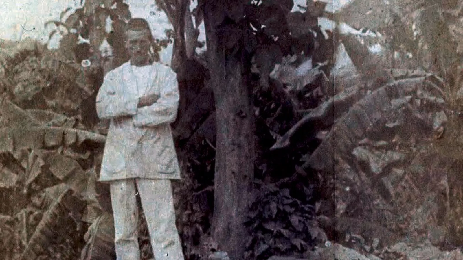 Fotografia antiga e desgastada, em tons de sépia, de Arthur Rimbaud adulto, de pé ao lado de uma árvore, com os braços cruzados. Ele veste terno claro e está cercado por folhagens densas, como folhas de bananeira.