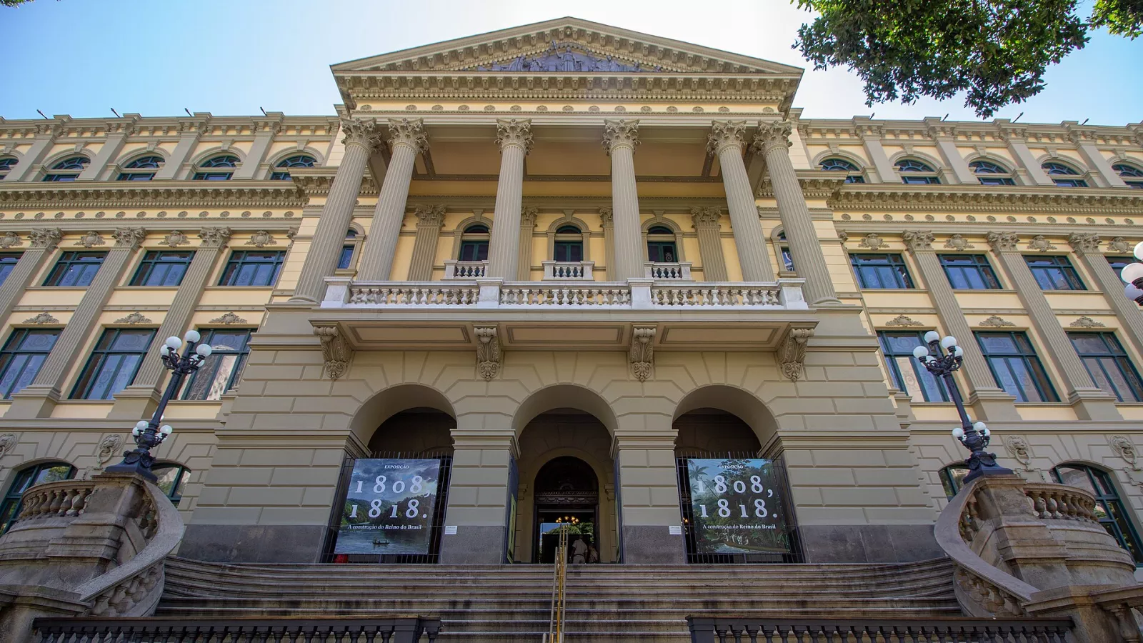 Fachada da Biblioteca Nacional, no Rio de Janeiro, vista de baixo para cima num dia de céu azul. O prédio é amarelo-claro, em estilo eclético, com colunas coríntias, frontão triangular, sacada com balaustrada e três arcos na entrada. Uma escadaria leva até a porta, ladeada por postes de luminárias. Dois painéis nos arcos laterais anunciam a exposição "1808, 1818. A construção do Reino do Brasil".