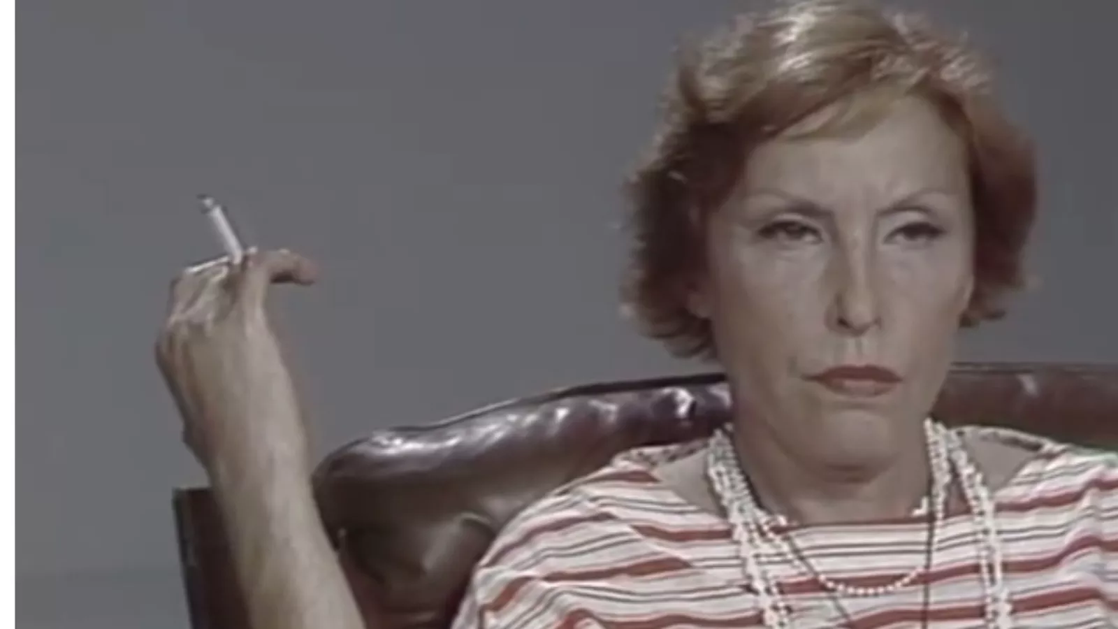 Clarice Lispector, de cabelo ruivo curto e colares de contas brancas, sentada numa poltrona de couro marrom diante de um fundo cinza, segurando um cigarro e olhando para a câmera com expressão séria, em imagem de vídeo.
