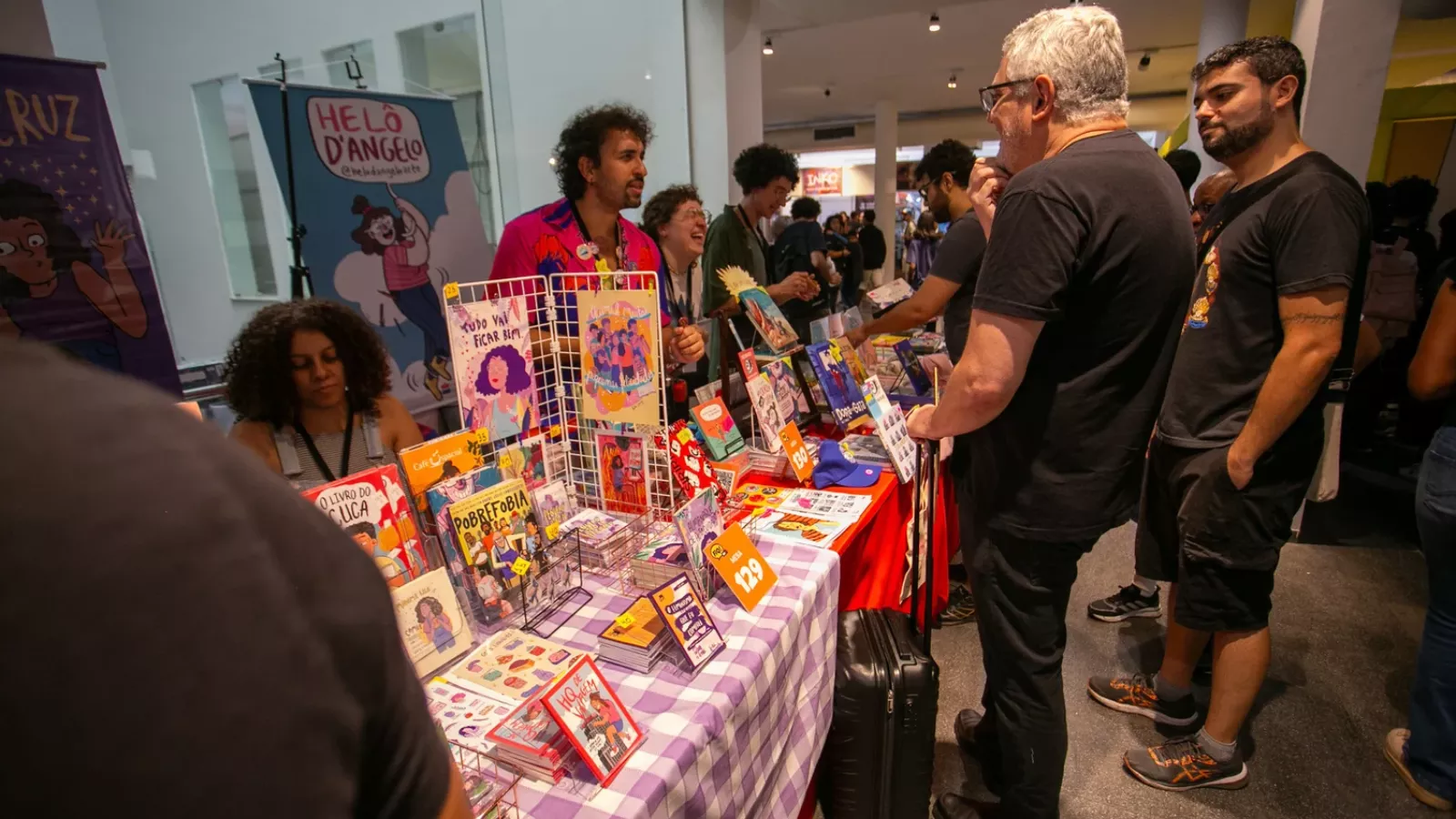 Mesa de quadrinistas numa feira de quadrinhos, coberta com toalha xadrez lilás e cheia de HQs e zines à venda. Atrás da mesa, um banner da quadrinista Helô D’Angelo. Visitantes conversam com os autores, e um deles puxa uma mala de rodinhas.