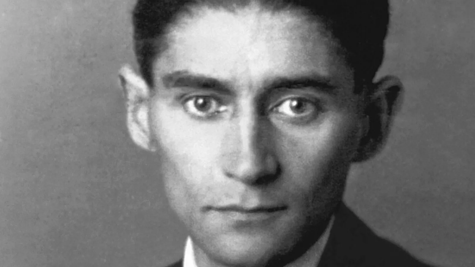Fotografia em preto e branco de Franz Kafka, em close, olhando diretamente para a câmera. Ele tem cabelo escuro e curto penteado para trás, sobrancelhas grossas, olhos escuros e orelhas salientes, e veste paletó escuro com camisa branca.
