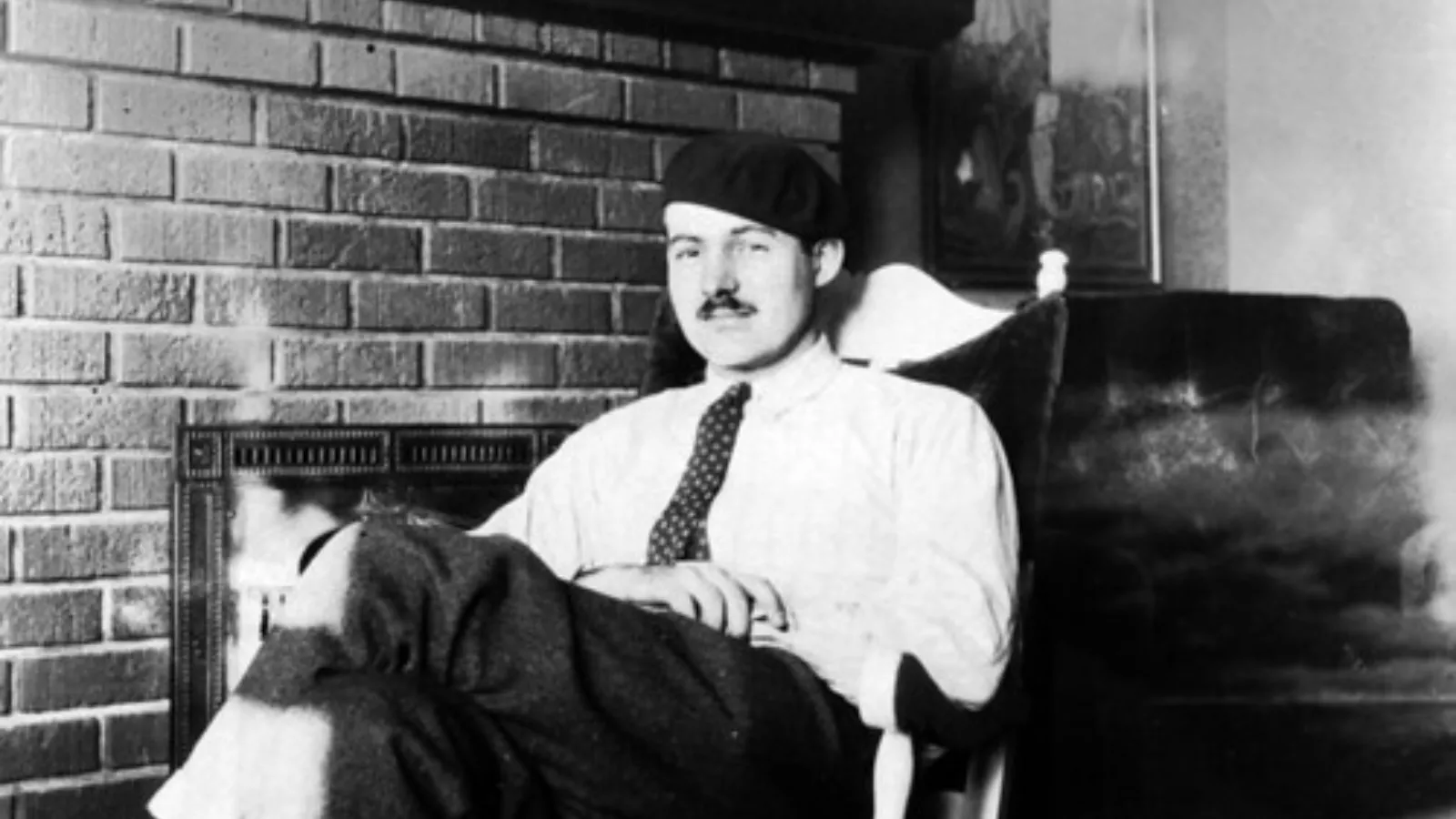 Ernest Hemingway jovem sentado em uma poltrona, usando camisa, gravata e um gorro escuro.