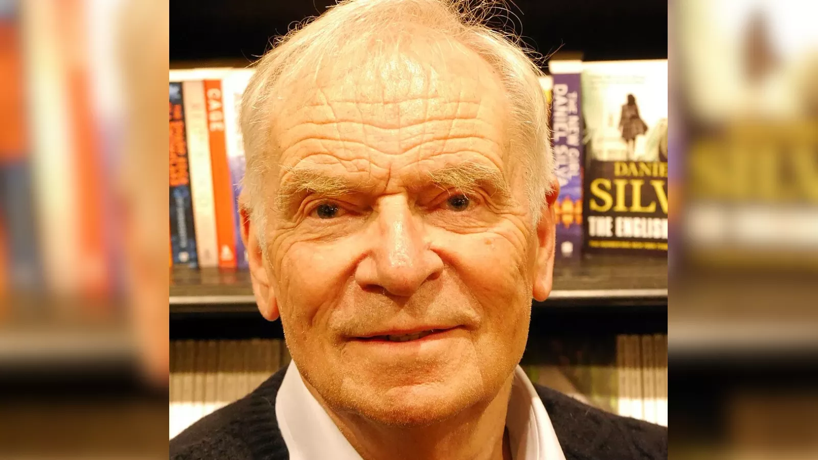 Jeffrey Archer sorridente, de cabelos brancos, camisa clara e suéter escuro, diante de uma estante de livros na livraria Hatchards, em Londres, em 2024, escritor britânico de Kane e Abel