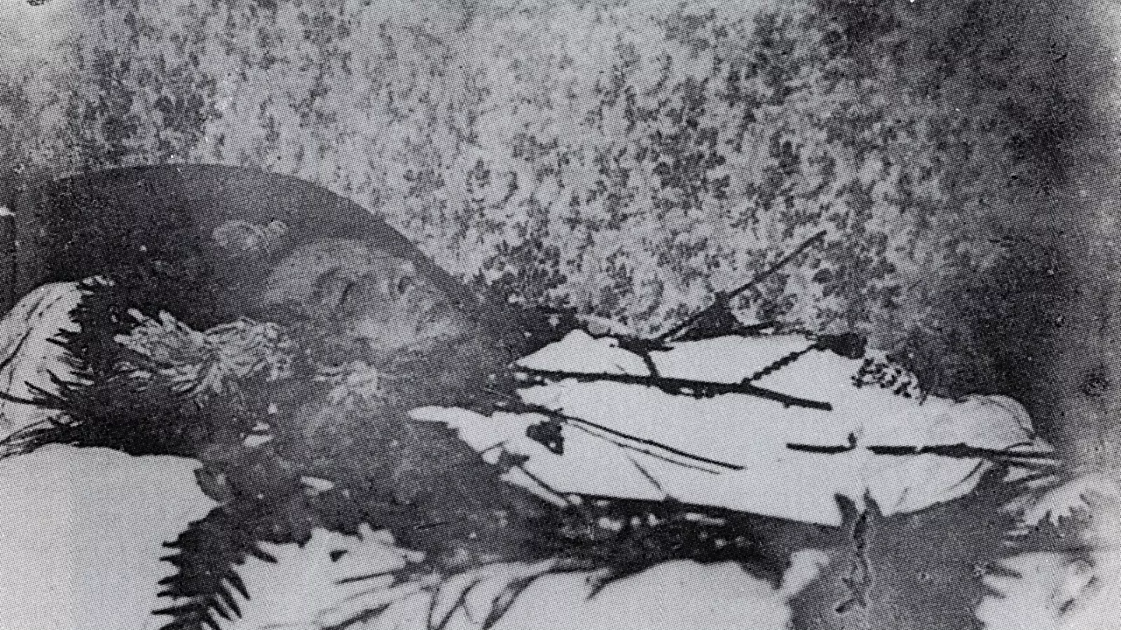 Fotografia antiga em preto e branco, granulada e de baixa definição, de Oscar Wilde morto, deitado de perfil numa cama. Há folhagens e flores sobre o lençol e um pequeno objeto pousado sobre o corpo.