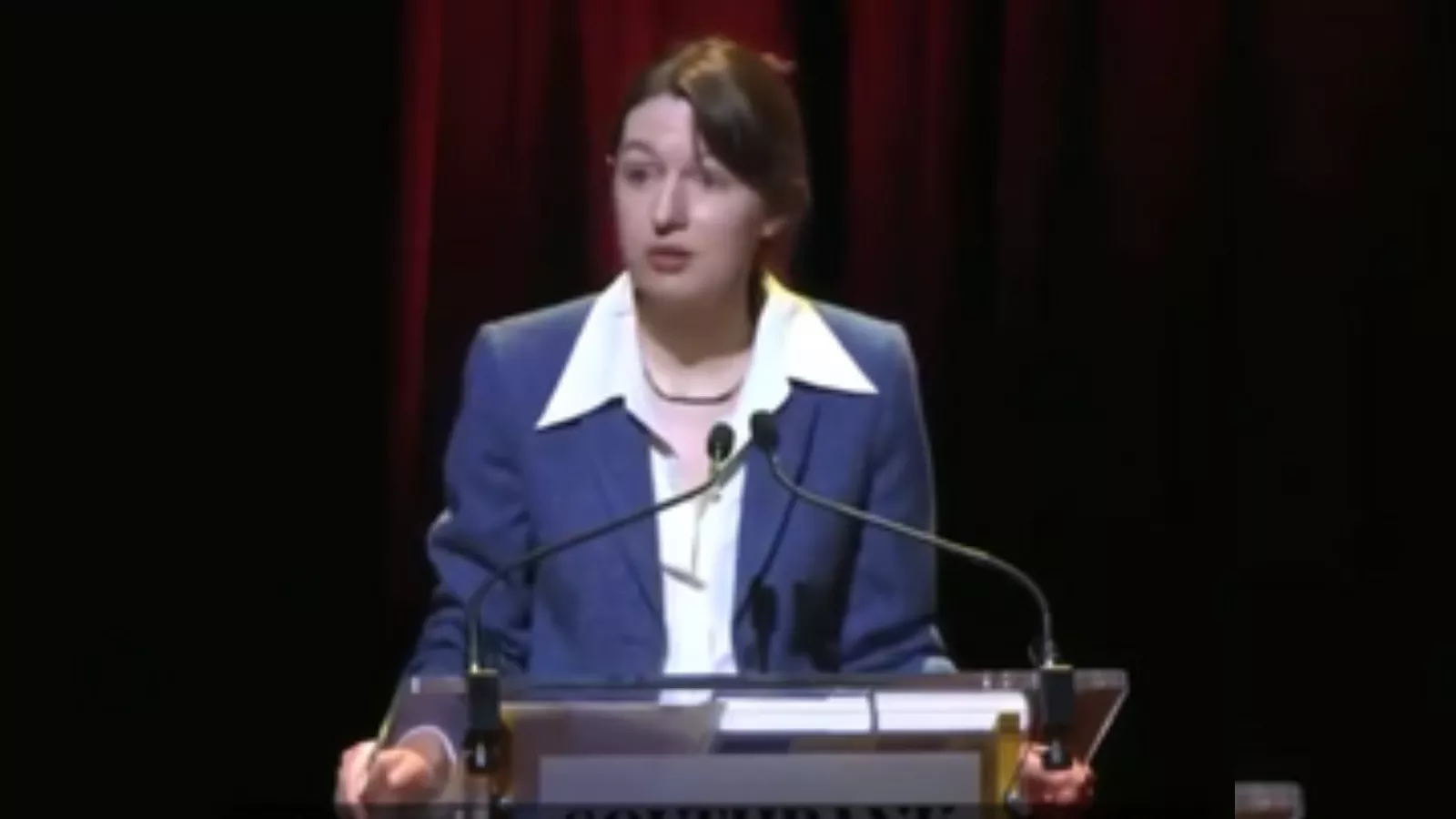 Sally Rooney discursa num púlpito transparente com dois microfones. Ela usa blazer azul sobre camisa branca de gola larga e tem o cabelo castanho preso, diante de uma cortina vermelha num palco escuro.