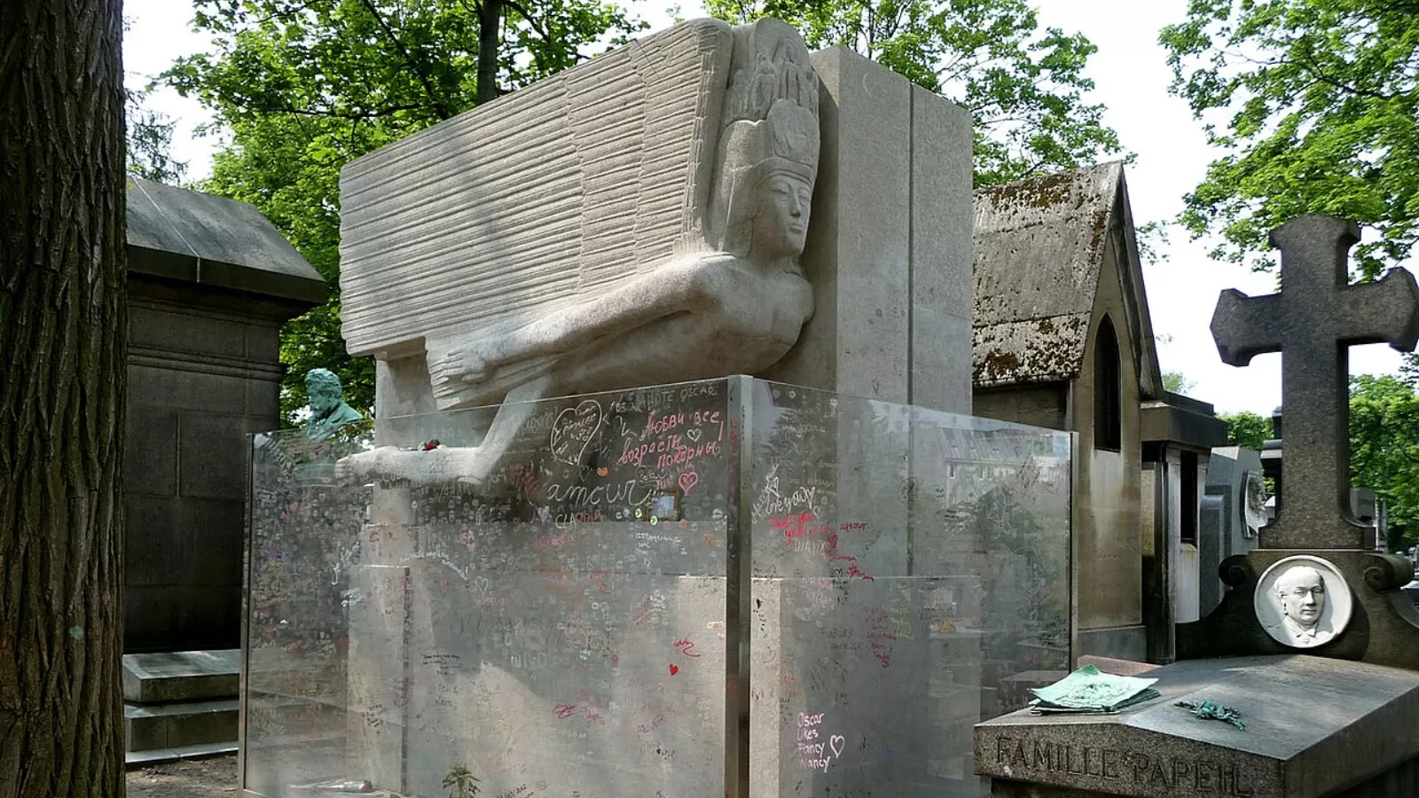 Túmulo de Oscar Wilde no cemitério do Père-Lachaise, em Paris. O monumento de pedra clara traz, em relevo, uma figura alada e esguia, de rosto estilizado e coroa alta, projetada para a frente. A base está cercada por painéis de vidro cobertos de recados, corações e palavras escritos à mão, entre eles "amour". Em volta há árvores, jazigos antigos e uma cruz de pedra.