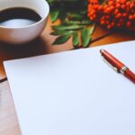 Uma página em branco com uma caneta, uma xícara de café e uma rosa acima na mesa