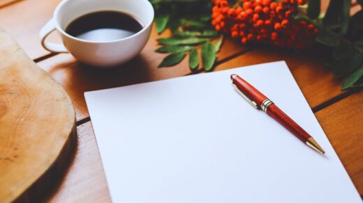 Uma página em branco com uma caneta, uma xícara de café e uma rosa acima na mesa