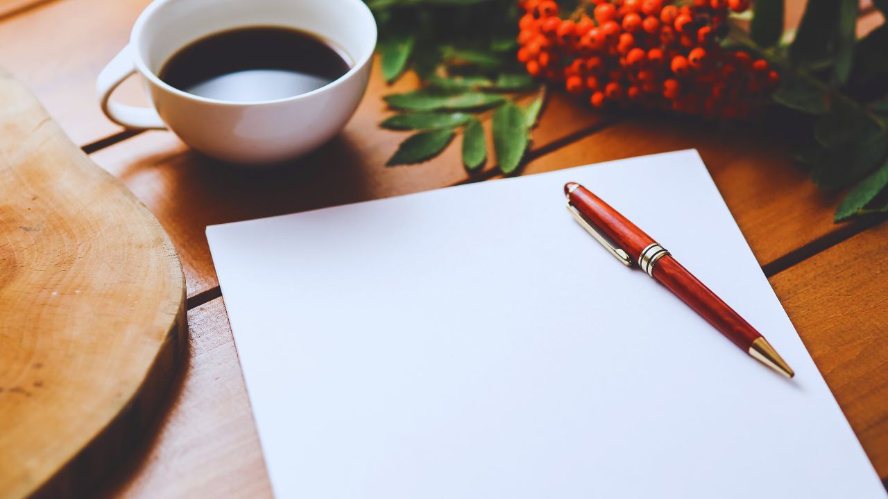 Uma página em branco com uma caneta, uma xícara de café e uma rosa acima na mesa