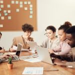 Um grupo diverso de colegas trabalhando em laptops e analisando documentos em uma reunião de equipe, com um quadro de anotações ao fundo