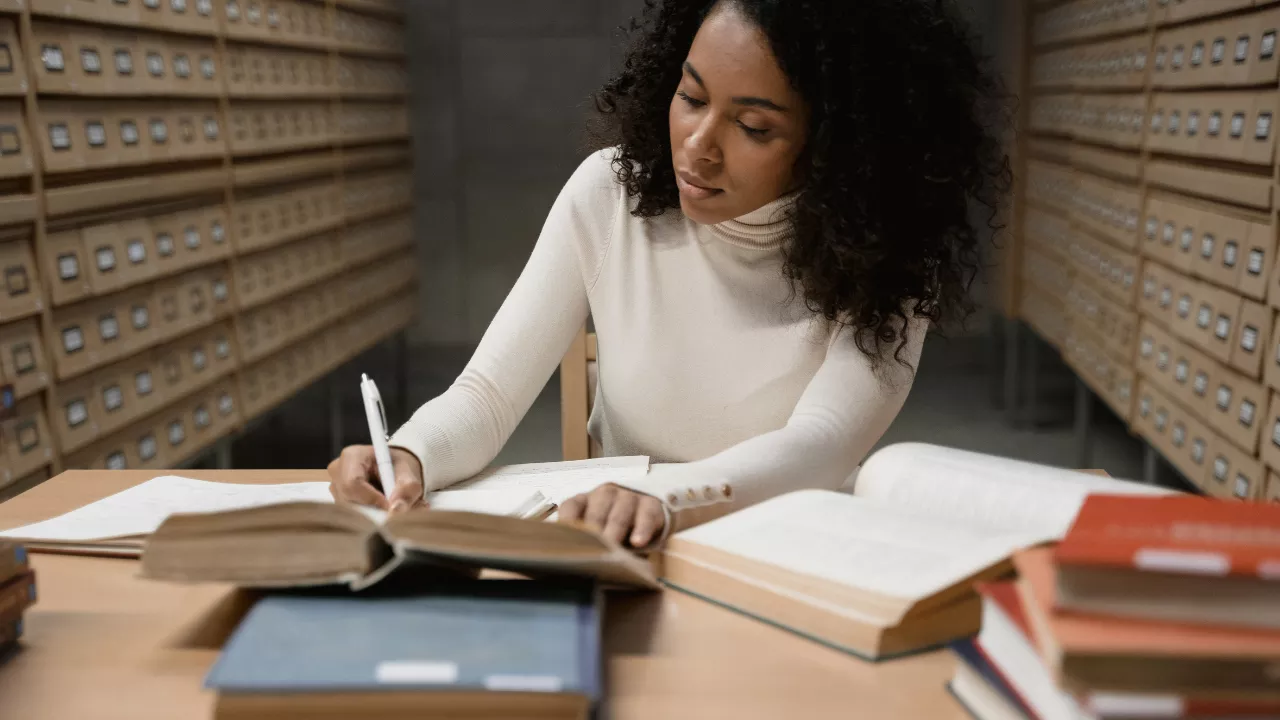 Mulher estudiosa escrevendo em um caderno, cercada por livros em uma biblioteca. Ilustra o aprofundamento da pesquisa acadêmica, essencial na elaboração de monografias, dissertações e teses.