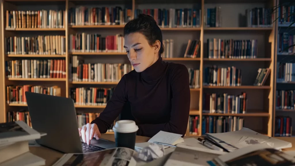 Jovem pesquisadora concentrada no laptop, rodeada de livros e anotações em uma biblioteca. Representa o processo de escrita e revisão de trabalhos acadêmicos em níveis avançados de estudo.