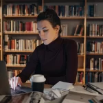 Jovem pesquisadora concentrada no laptop, rodeada de livros e anotações em uma biblioteca. Representa o processo de escrita e revisão de trabalhos acadêmicos em níveis avançados de estudo.