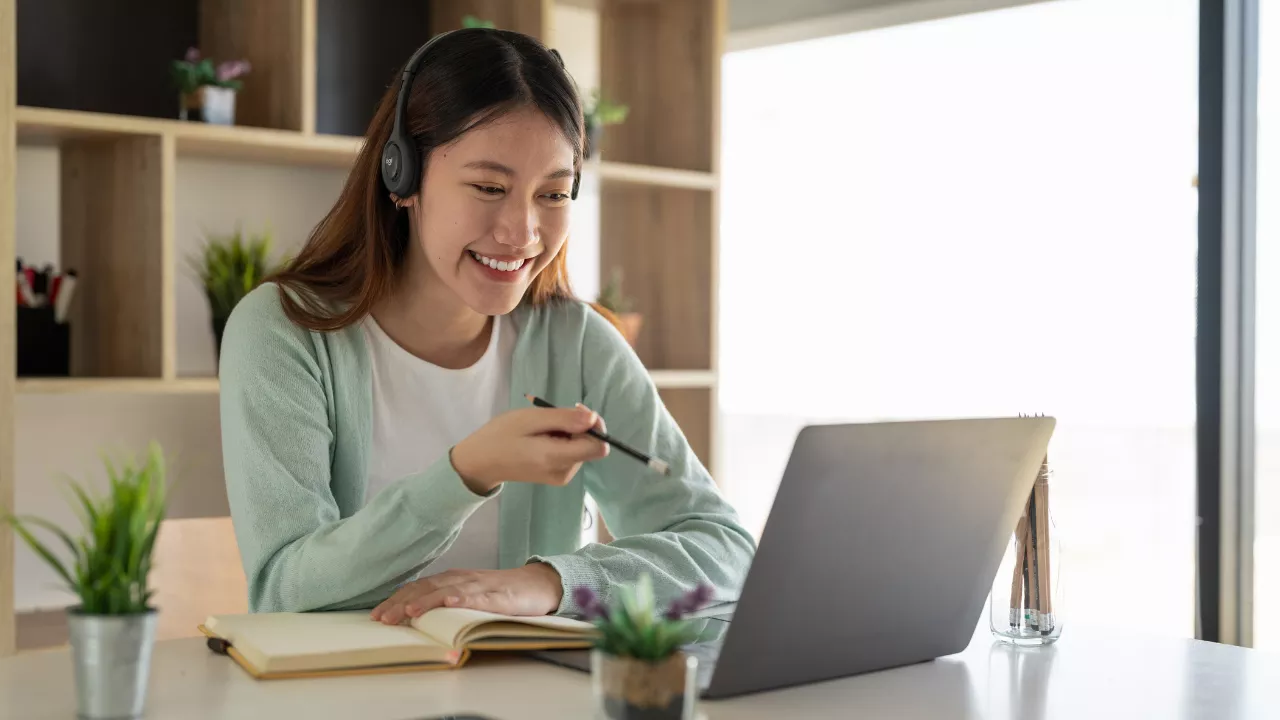 Jovem estudante com fones de ouvido sorrindo enquanto faz anotações e participa de uma aula online pelo laptop. Representa o processo de aprendizado e pesquisa acadêmica.
