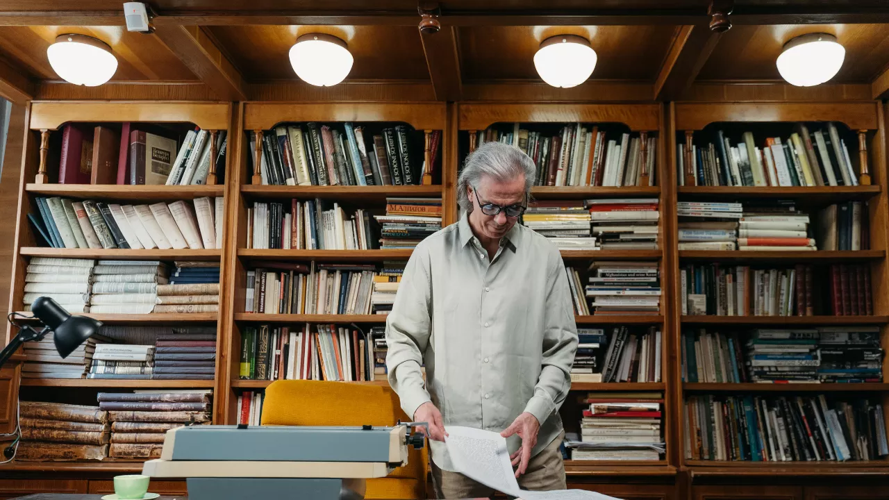 Homem mais velho em uma biblioteca clássica, revisando documentos ao lado de uma máquina de escrever. Simboliza a experiência acadêmica e a produção de conhecimento avançado, como dissertações e teses.