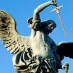 Estátua do Arcanjo Miguel com asas abertas e espada levantada contra um céu azul intenso. A figura, de aparência imponente, retrata São Miguel em posição de combate, simbolizando sua força espiritual como defensor contra o mal. A escultura representa a imagem clássica do príncipe da milícia celeste, reforçando visualmente os temas de proteção, fé e justiça abordados na oração de São Miguel Arcanjo.