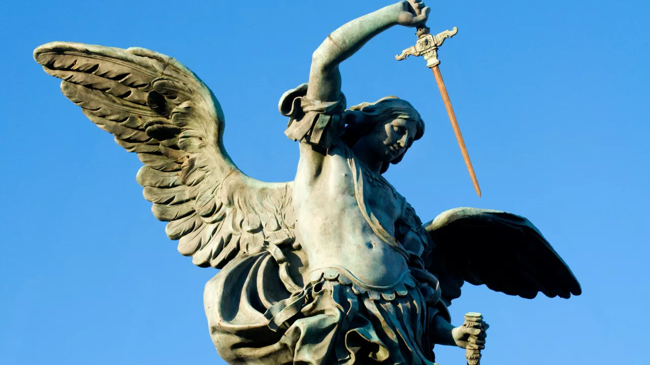 Estátua do Arcanjo Miguel com asas abertas e espada levantada contra um céu azul intenso. A figura, de aparência imponente, retrata São Miguel em posição de combate, simbolizando sua força espiritual como defensor contra o mal. A escultura representa a imagem clássica do príncipe da milícia celeste, reforçando visualmente os temas de proteção, fé e justiça abordados na oração de São Miguel Arcanjo.