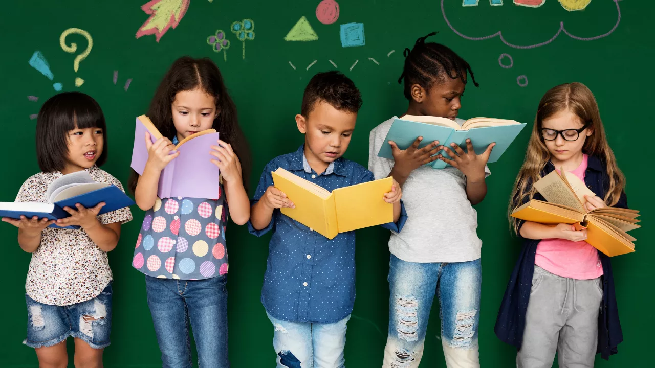 Grupo de crianças pequenas lendo livros coloridos diante de um quadro verde decorado com desenhos infantis. A cena expressa curiosidade, aprendizado e diversidade, reforçando a importância de oferecer referências plurais e representativas no ambiente escolar.
