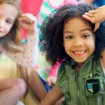 Crianças sorridentes brincam juntas, com destaque para uma menina negra de cabelo crespo em primeiro plano, expressando alegria e autoconfiança. A imagem reflete o impacto positivo de práticas pedagógicas que valorizam a identidade e fortalecem a autoestima desde a Educação Infantil.